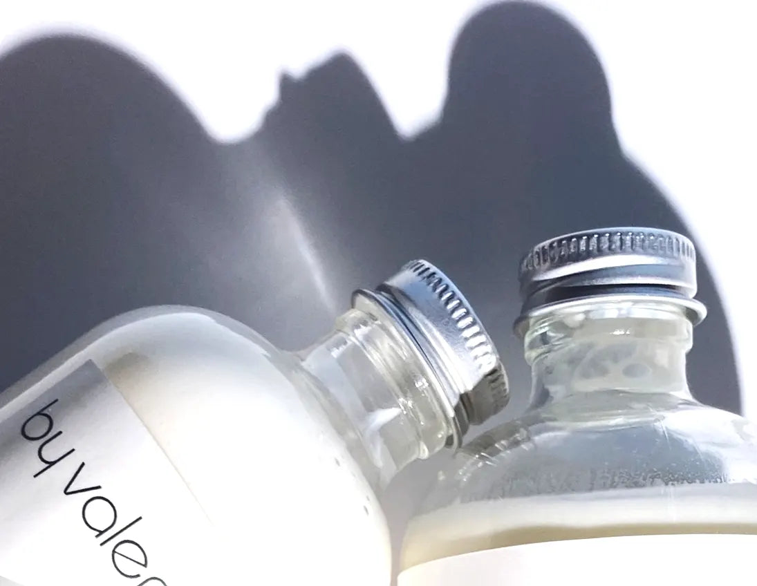 By Valenti close up of glass bottles of body lotion on a white background casting a long afternoon shadow