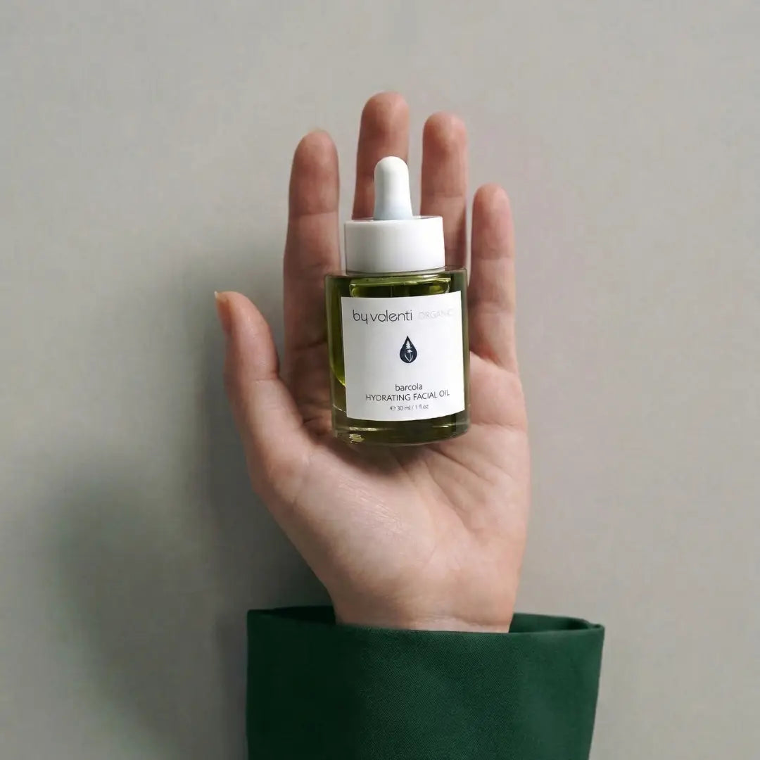 Hand holding a glass bottle with dropper of By Valenti Barcola Hydrating Facial Oil with a neutral background. Model is wearing a green sleeve matching the green colored serum in the transparent bottle.