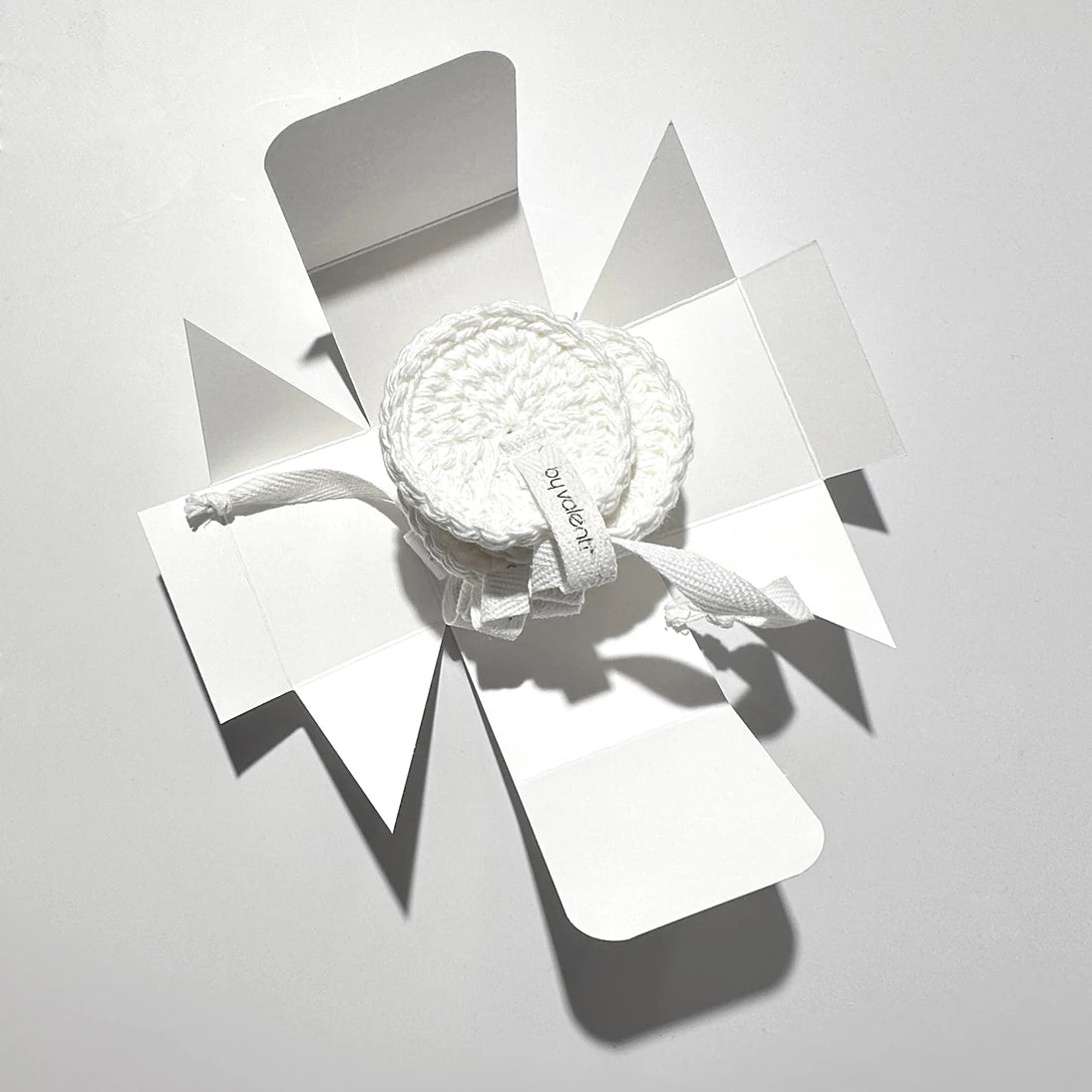 By Valenti white crocheted facial cotton rounds or pads in a deconstructed white box on a light gray background