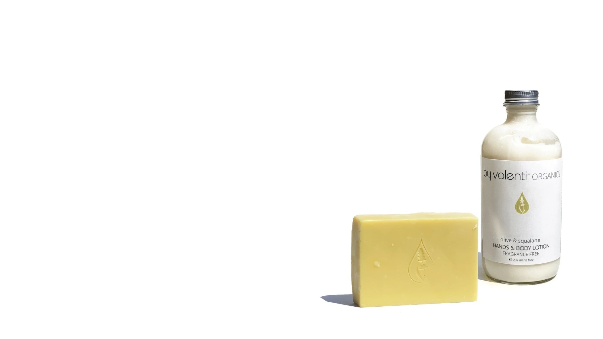 A clear glass bottle filled with By Valenti golden-amber Castile Liquid Soap and Body Wash and a True Castile soap bar, both with white labels, photographed on a white background with a hard shadow.