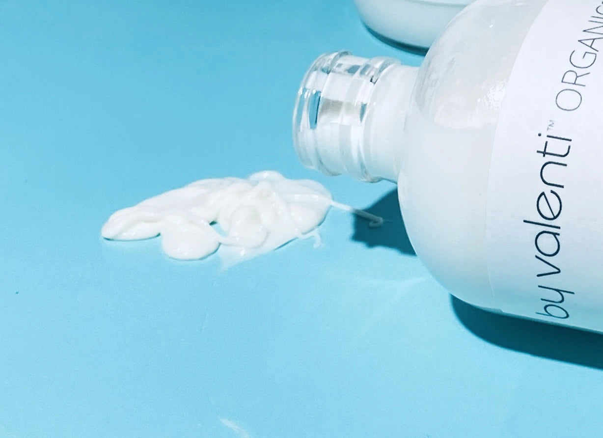 By Valenti Organics glass bottle of body lotion spilling on a blue background