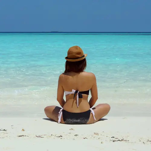 Woman wearing a black and white bikini and a hat sitting on a beach with clear blue water and sky