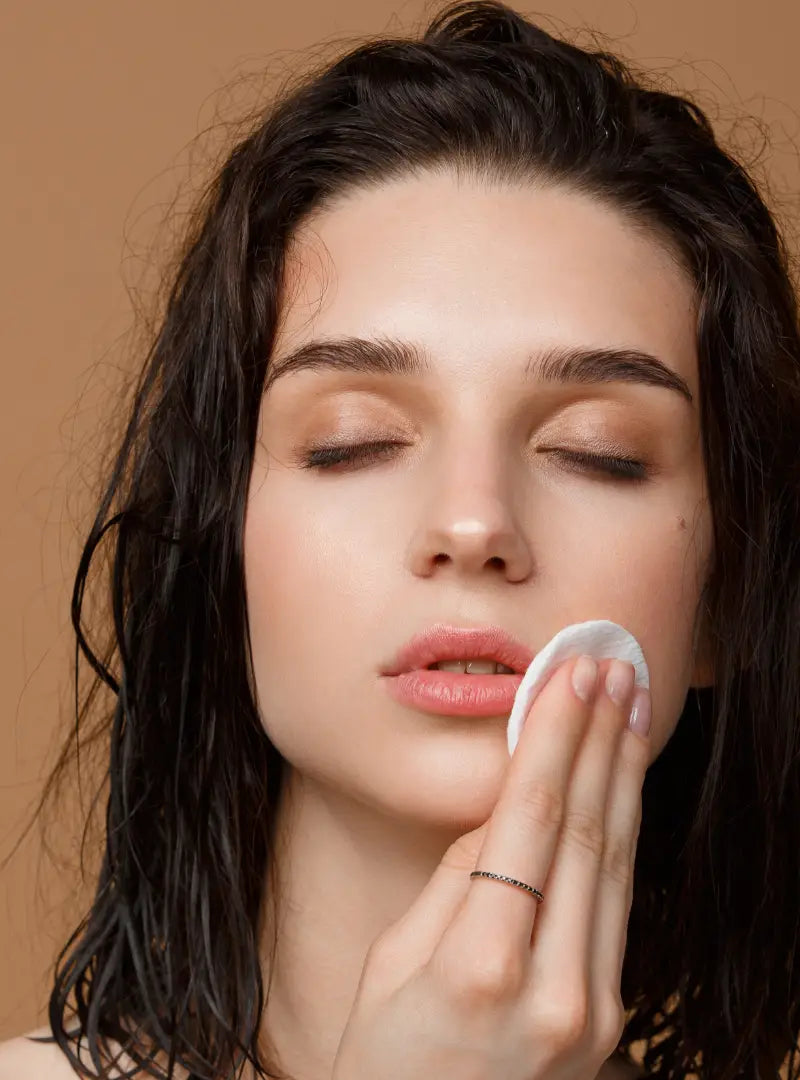 Woman applying a product to her face with a cotton pad against a neutral background
